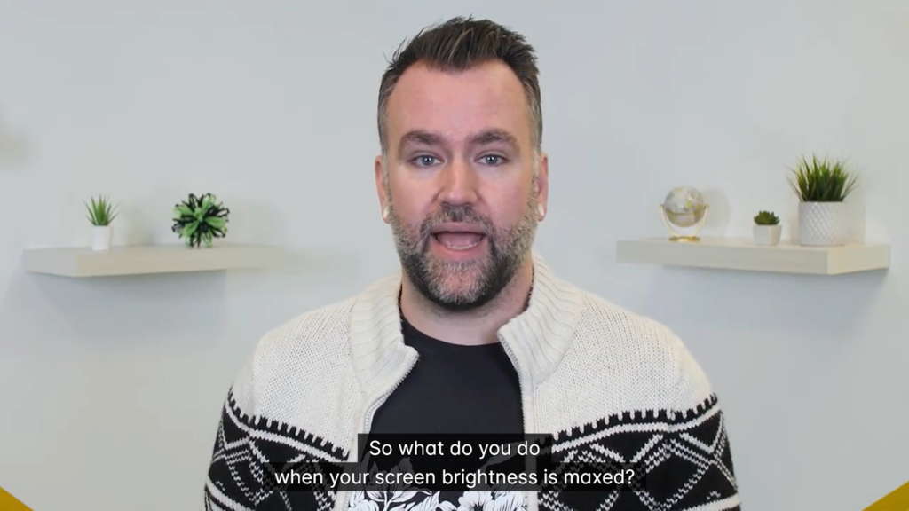 A man is speaking in front of a camera. The captions read "So what do you do when your screen brightness is maxed?" The captions are on two lines and have a backdrop for contrast.