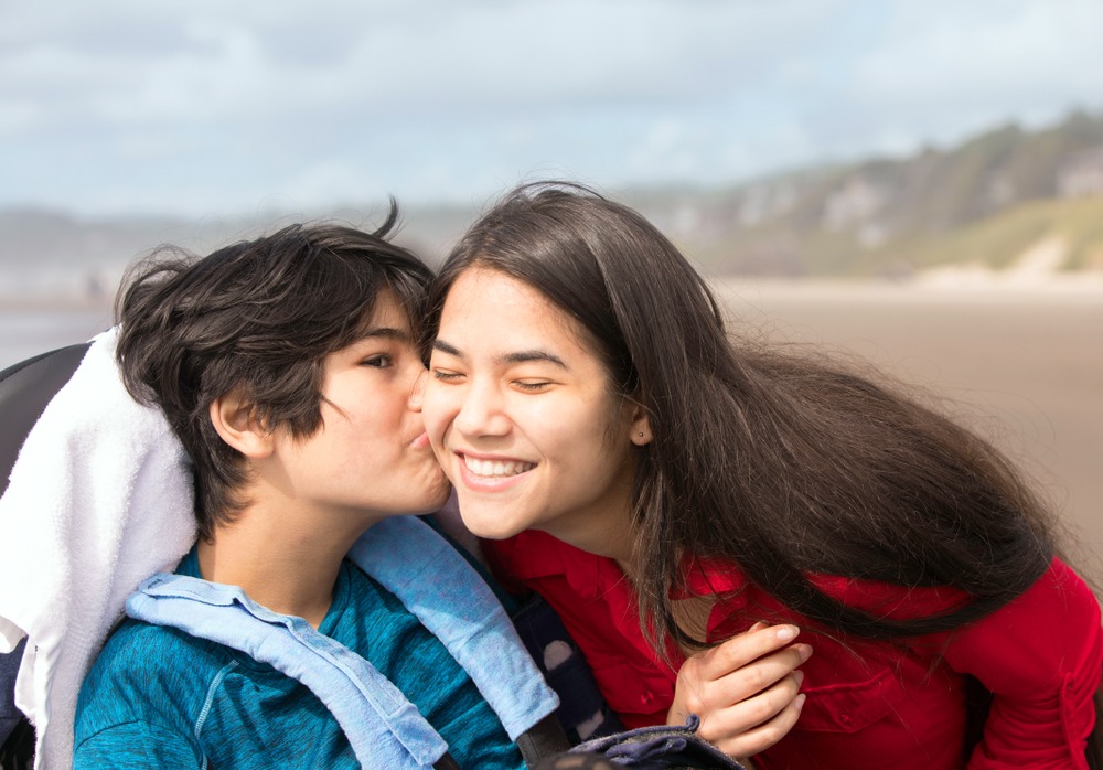 A child in a wheelchair is kissing a woman on the cheek. The woman is smiling brightly.